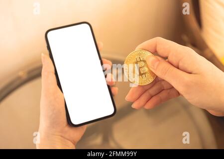 Pagamenti con un mockup di valuta criptata, telefono cellulare con schermo bianco vuoto e mano donna con moneta di bitcoin oro, spazio di copia Foto Stock