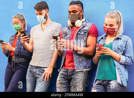 Gruppo di persone che utilizzano il loro smartphone in covid 19 volte protetto con maschera viso - amici che controllano le notizie online mentre si levano in piedi al muro e holdi Foto Stock