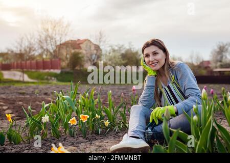 Sorridente giardiniere rilassante tra tulipani freschi, narcisi, giacinti nel giardino primaverile. Donna felice ammira fiori colorati seduti a terra Foto Stock