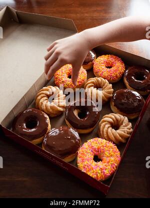 Mano del bambino che raggiunge in una scatola di ciambelle assortite su un tavolo. Foto Stock