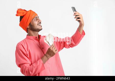 Giovane agricoltore indiano utilizzando smartphone e mostrando denaro su sfondo bianco. Foto Stock