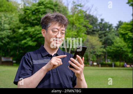 Uomo asiatico di mezza età che usa uno smartphone sul prato. Foto Stock