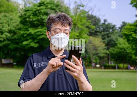 Un uomo asiatico di mezza età che indossa una maschera facciale e utilizza uno smartphone sul prato. Foto Stock