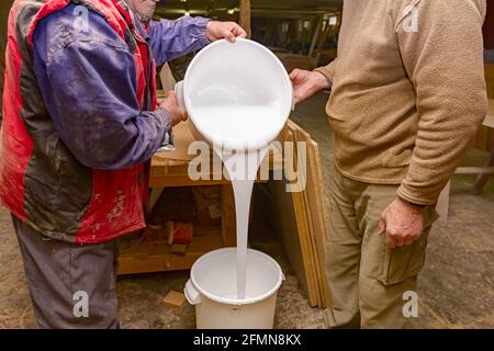 Il lavoro di squadra di due falegnami sta versando colla per incollare legno da un secchio di plastica ad un altro che è vuoto. Foto Stock