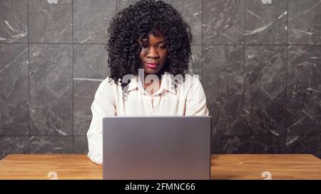 Attraente addetta afroamericana di affari in vestiti eleganti siede giù a. lavora su un computer portatile moderno al tavolo vicino alla parete grigia ufficio Foto Stock