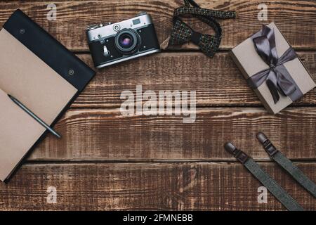 Confezione regalo per il giorno del padre con gli accessori da uomo con cravatta a prua, portafoglio, fotocamera retrò, sospensori e scarpe in pelle e blocco note su sfondo in legno. Copia Foto Stock