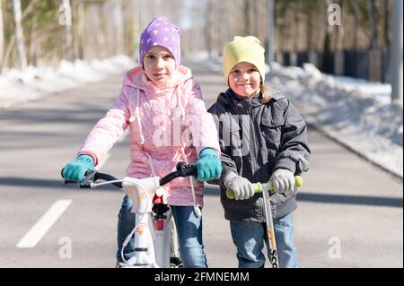 Mosca, Russia, marzo 2021 - felici fratelli e amici, un fratello minore su uno scooter e una sorella ragazza su una bicicletta nel parco della città. saf Foto Stock