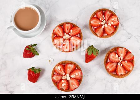 Vista dall'alto di piccoli pasticcini al tartlet ricoperti di fragole e il cioccolato bianco spruzzerà accanto alla tazza di caffè Foto Stock