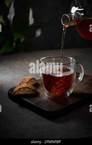 Il tè nero viene versato dalla teiera in una tazza trasparente con bolle. Biscotti nelle vicinanze sul tagliere. Messa a fuoco selettiva. Orientamento verticale, tasto basso, rustico Foto Stock