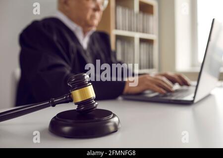 Primo piano di gavel marrone scuro sul tavolo sullo sfondo di un giudice maschile senior che lavora su un computer portatile. Foto Stock