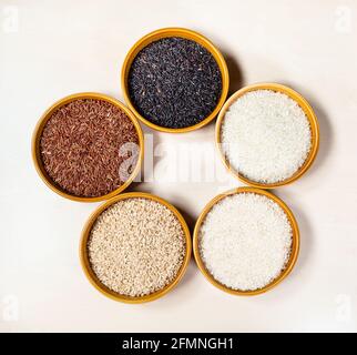 vista dall'alto di vari grani di riso (nero, rosso, bromo, lucidato, non lucidato, bianco) in ciotole rotonde in ceramica su tavola di legno marrone chiaro Foto Stock