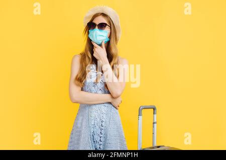 Pensando giovane donna viaggiatore, indossando una maschera medica protettiva, indossando un cappello, con una valigia su sfondo giallo, i passeggeri che viaggiano all'estero Foto Stock