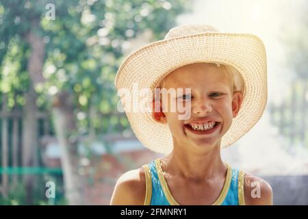 Ragazzo allegro che indossa un grande cappello di paglia e senza maniche blu una camicia che si posa per la macchina fotografica e sorride in bocca nel soleggiato parco cittadino estivo vicino Foto Stock