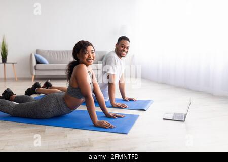 Una coppia nera sportiva che fa il backbend sui tappetini yoga, seguendo il tutorial online sul computer portatile in interni, spazio vuoto Foto Stock