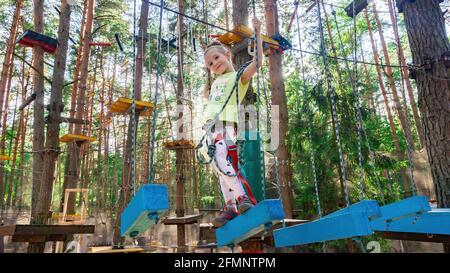 Bella ragazza che si diverte nel parco avventura corda. Una ragazza in età scolare cammina lungo blocchi di legno sospesi da una corda e sorride. Foto Stock