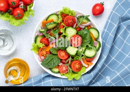 Insalata di verdure estive con pomodori ciliegini, cetrioli, rucola, basilico e condita con olio d'oliva. Tovagliolo a scacchi blu su un tavolo grigio. Vista da a. Foto Stock
