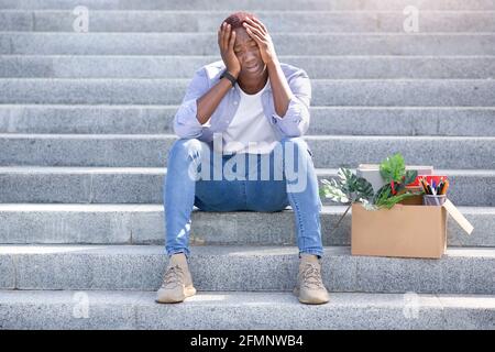 Crisi economica. Disperato uomo afro-americano con oggetti personali seduti sulle scale all'aperto dopo essere stato licenziato Foto Stock