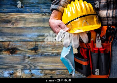 Elettricista lavoratore su sfondo di legno blu chiaro vintage; tiene casco, guanti, occhiali e la maschera chirurgica per prevenire la diffusione di Coronavirus. Foto Stock