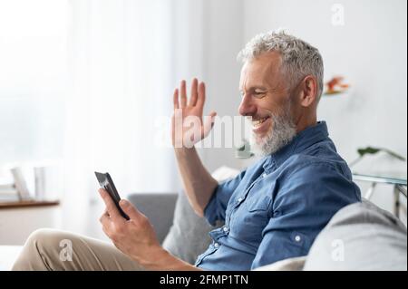 Concetto di videochiamata. Divertente hipster uomo di mezza età con capelli grigi in camicia casual con smartphone per riunioni virtuali seduto sul divano di casa, un ragazzo maturo ama chattare online e fare un saluto Foto Stock
