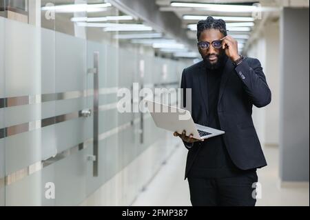 Ritratto di un afroamericano in abito nero sullo sfondo dell'ufficio. Uomo d'affari nero con un computer portatile Foto Stock