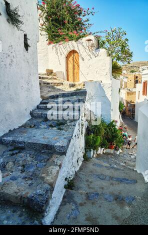 Strette strade ripide con case bianche pareti e cespugli fioriti E vecchie scale nella storica città greca Lindos sotto il blu cielo Foto Stock