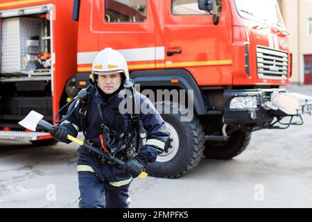 Ritratto di un serio e sicuro pompiere caucasico in piedi e che tiene il martello, indossando una speciale uniforme protettiva sullo sfondo del camion. Foto Stock