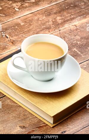 Tazza bianca e piattino ripieno di tè seduto su un libro di lettura su un tavolo rustico in legno Foto Stock