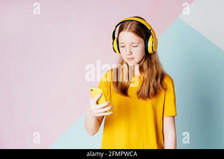 Ragazza hipster con capelli rossi in cuffie wireless ascolta una playlist su un telefono cellulare. Foto Stock