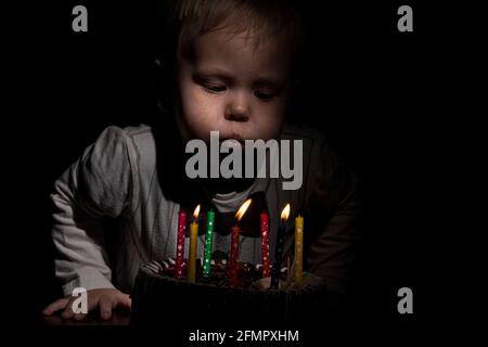 Compleanno. Un piccolo ragazzo dolce soffia le candele sulla stoca. Sfondo scuro. Foto Stock