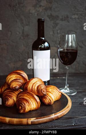 Croissant profumati appena sfornati su un pannello di pietra scura, un bicchiere di vino rosso e una bottiglia. Cena romantica. Foto Stock