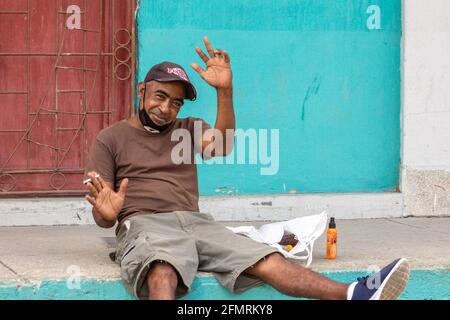 Ritratto candido di un cubano afro caraibico seduto sul marciapiede. Sta fumando così la sua maschera protettiva è giù. È il tempo del Covid Foto Stock