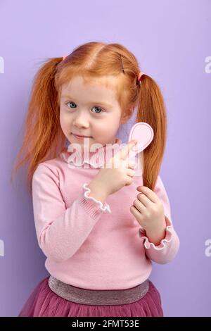 Bambina che combatte i capelli rossi che posano alla macchina fotografica, isolato sullo sfondo viola dello studio. Ritratto di adorabile carino bambino che prende cura della condizione dei capelli Foto Stock