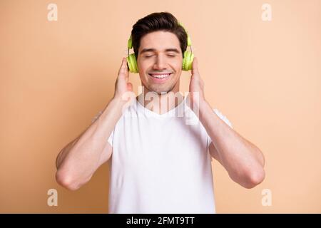 Foto di un giovane allegro uomo chiuso occhi braccia che toccano le cuffie isolato su sfondo di colore beige Foto Stock