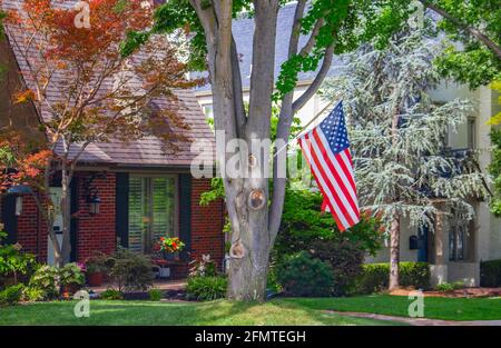 Casa in mattoni situata in un quartiere tradizionale con grandi alberi a. Alimentatore di uccelli e fiori colorati e una bandiera americana Foto Stock