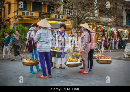 Marchette di frutta vietnamite che trasportano i cestini gemelli e le aste delle spalle che offrono ai turisti le loro merci per le occasioni fotografiche, Hoi An, Vietnam Foto Stock