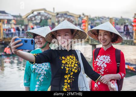 Tre Signore vietnamite che indossano cappelli conici e abiti tradizionali ao dai posano per selfie dal lungofiume, Hoi An, Vietnam Foto Stock