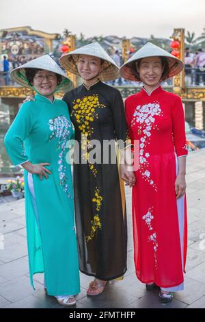 Tre Signore vietnamite che indossano abiti tradizionali di ao dai e cappelli conici posano vicino al fiume nella città vecchia, Hoi An, Vietnam Foto Stock