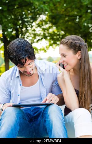 L uomo e la donna o una giovane coppia di bere il caffè e seduta nel parco della città in estate e utilizzo di smart phone e computer tablet Foto Stock