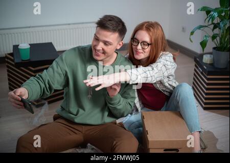 Una ragazza carina e un ragazzo mostrano le chiavi del loro nuovo appartamento nel loro smartphone e sono felici con l'acquisto. Foto Stock