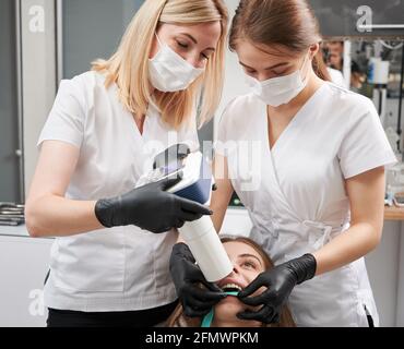 Dentista femminile e il suo assistente che fa l'immagine a raggi X con una macchina a raggi X portatile. Persone che indossano uniforme clinica, maschere per il viso e guanti neri. Concetto di odontoiatria digitale e radiografia intraorale. Foto Stock