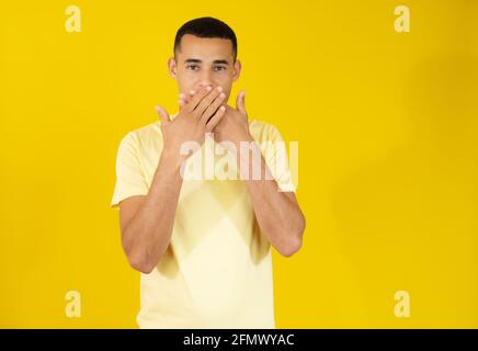 Giovane uomo in t-shirt casual che copre la bocca con le mani isolate su sfondo giallo. Foto Stock
