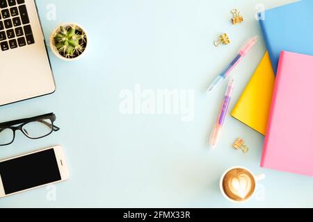 Luogo di lavoro in ufficio con computer portatile, diversi notebook colorati, telefono cellulare a schermo vuoto, tazza bianca di caffè, forniture e cancelleria su sfondo blu Foto Stock