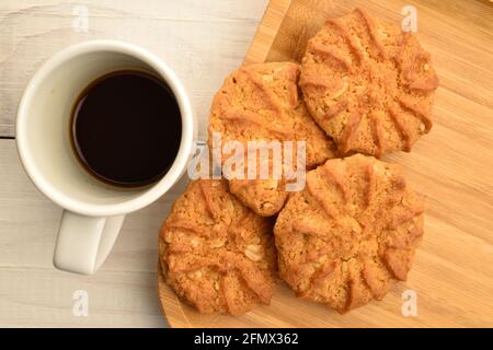 Diversi biscotti di cereali, fragranti rotondi, si trovano su un vassoio di bambù, accanto è una tazza di caffè, su un tavolo di legno bianco, vista dall'alto. Foto Stock