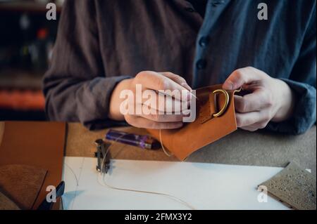 Seduto a un tavolo in officina, un ragazzo cucce manualmente gli elementi in pelle l'uno all'altro. Foto Stock