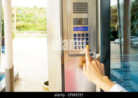 Primo piano della mano della donna che immette il codice del sistema di sicurezza, premendo il pulsante con il dito indice sul moderno dispositivo intercom con schermo lcd blu vicino all'entrata d Foto Stock