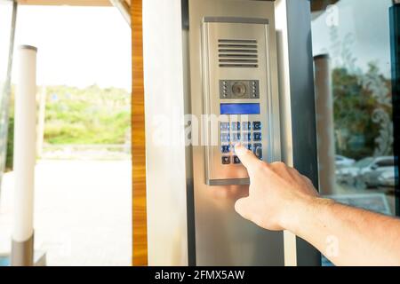 Primo piano della mano dell'uomo che immette il codice del sistema di sicurezza, premendo il tasto con il dito indice sul moderno dispositivo intercom con schermo lcd blu vicino entrence doo Foto Stock