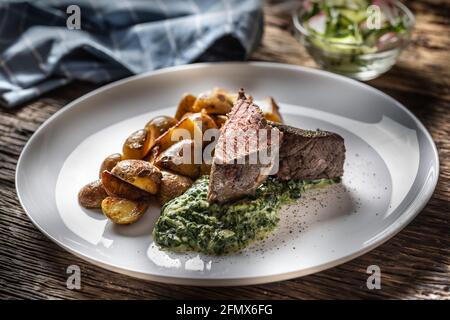 Deliziosa bistecca di manzo alla griglia, servita su un piatto bianco con patate arrosto croccanti, salsa di spinaci e insalata di lattuga a foglia fresca. Foto Stock