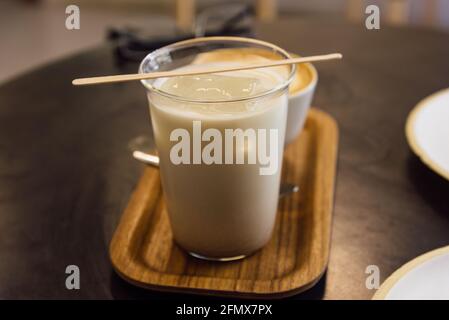 Latte di tè tailandese ghiacciato in tazza di vetro e cappuccino con vassoio di legno su tavolo di legno in una caffetteria. Foto Stock