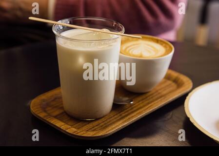 Latte di tè tailandese ghiacciato in tazza di vetro e cappuccino con vassoio di legno su tavolo di legno in una caffetteria. Foto Stock