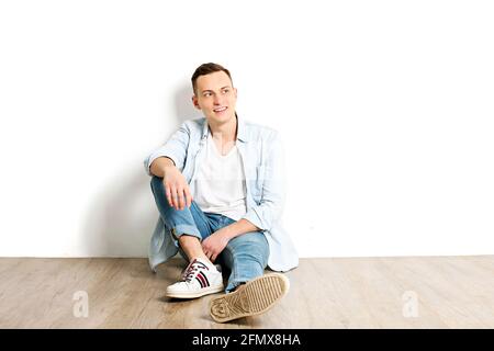 Ritratto di felice giovane uomo millenario con sorriso beaming in camicia bianca sbottonata si siede sul pavimento di tessitura di legno, isolato. Hippe casual sicuro di sé Foto Stock